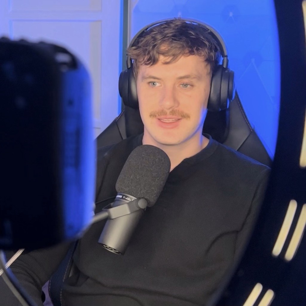 Ross lightly smiling in an office with a mic near his chest. He wears a pair of headphones and colorful blue lights can be seen in the background.